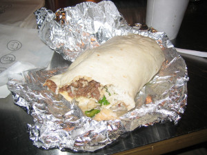 An image of a Chipotle burrito with the top eaten into. The burrito is surrounded by foil, napkins, and a drink.